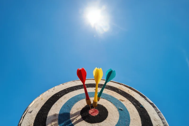 Dartboard with three colorful darts hitting the bullseye against a bright blue sky