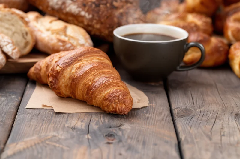 Freshly baked croissant on parchment paper beside a steaming cup of coffee on a rustic wooden table with assorted pastries in background