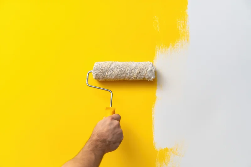 Hand holding paint roller applying white paint to a yellow wall during home renovation