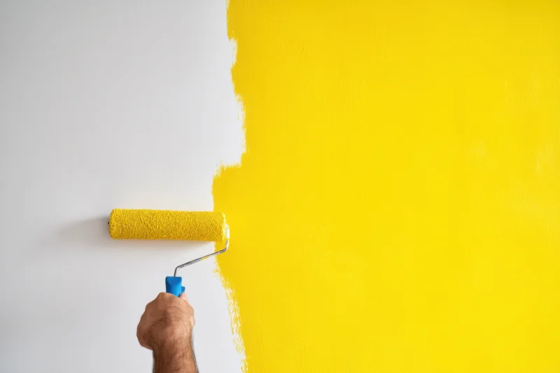 Hand using paint roller to apply yellow paint on white wall during home renovation