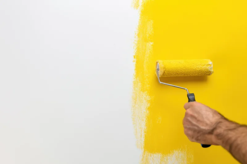 Hand holding paint roller applying yellow paint to white wall during home renovation