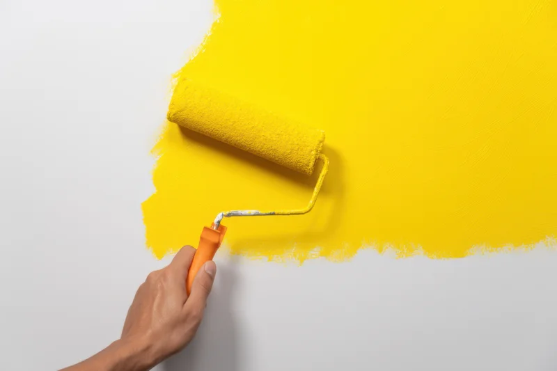 Hand using paint roller to apply yellow paint on white wall