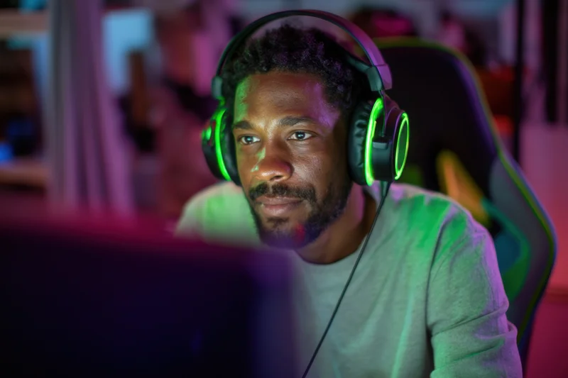 Black man focused on computer screen wearing glowing headset in dimly lit room