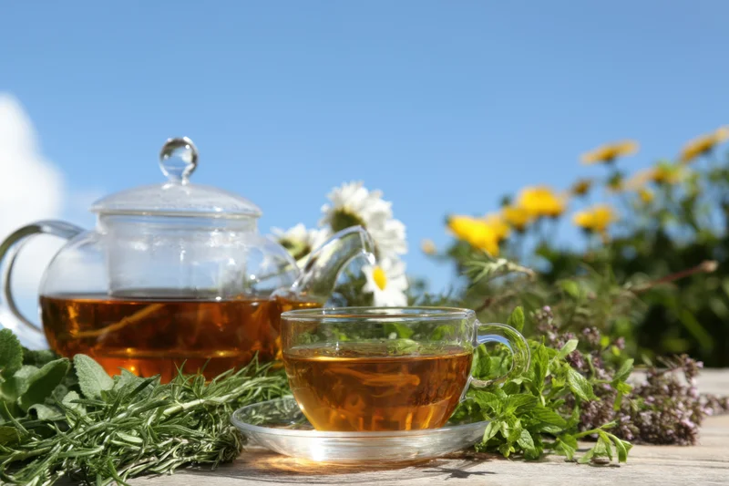 Glass teapot and teacup filled with amber herbal tea surrounded by fresh herbs and wildflowers on a wooden table outdoors