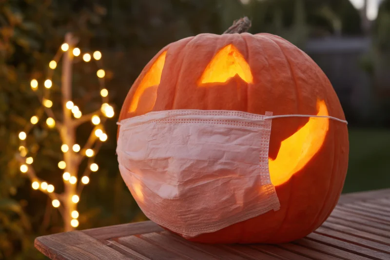 Carved pumpkin wearing a face mask glowing on wooden table with string lights in background