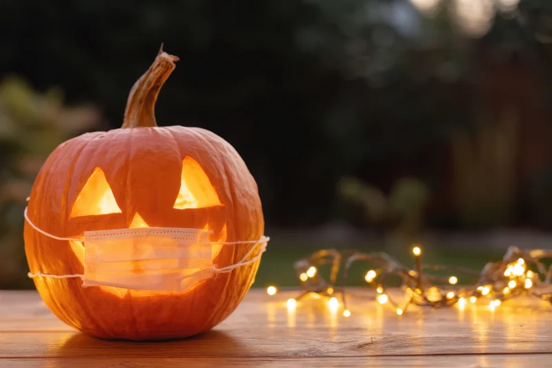 Carved pumpkin with glowing face wearing protective mask on wooden table near string lights
