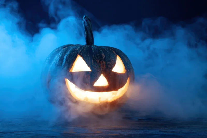 Glowing carved pumpkin with triangular eyes and wide grin surrounded by swirling blue mist on dark wooden surface at night