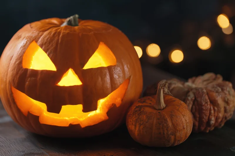Glowing jack-o-lantern with carved spooky face on wooden table beside smaller pumpkins and bokeh lights
