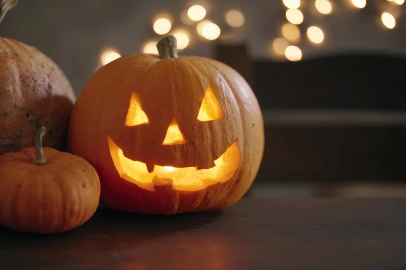 Glowing jack-o-lantern with carved spooky face on wooden table near string lights