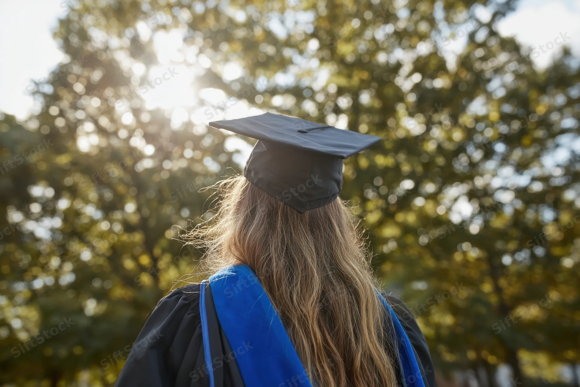 Graduate in cap and gown with blue sash standing outdoors facing trees in sunlight