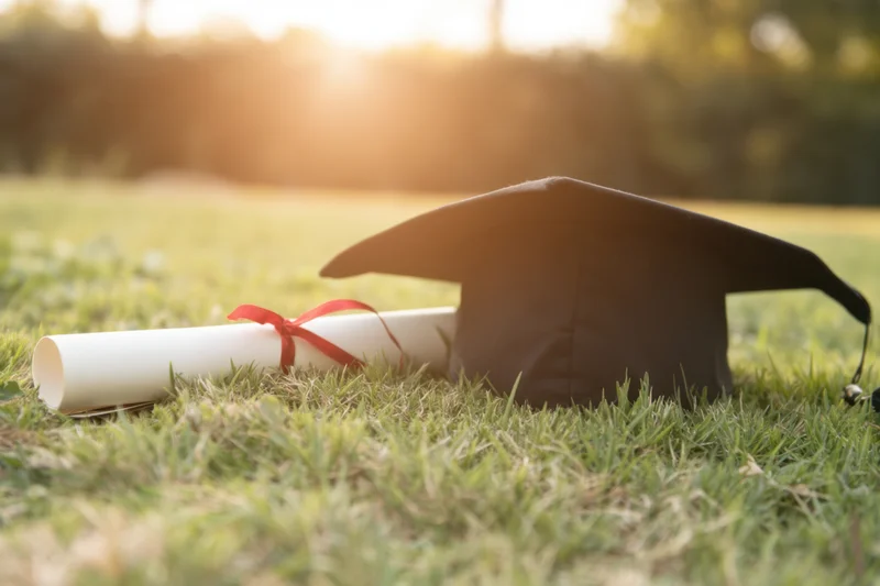 Graduation cap and rolled diploma with red ribbon resting on grassy field at golden hour