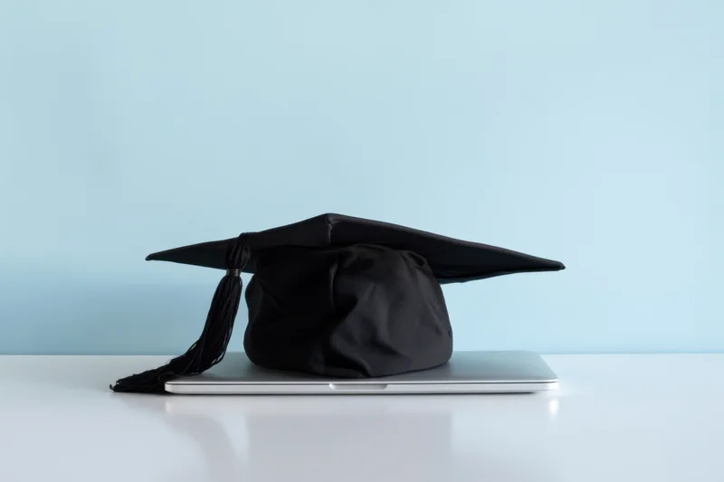 Graduation cap resting on closed laptop on white surface against blue background