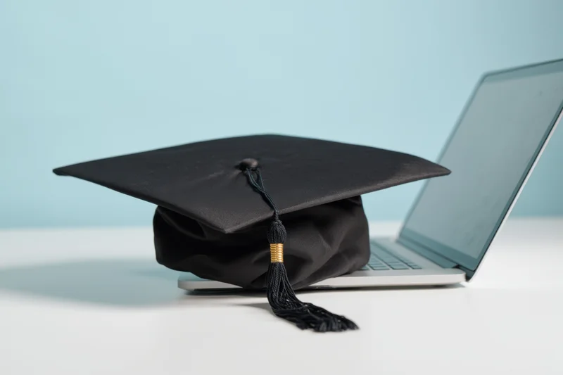 Graduation cap resting on a modern laptop on a white desk with blue background