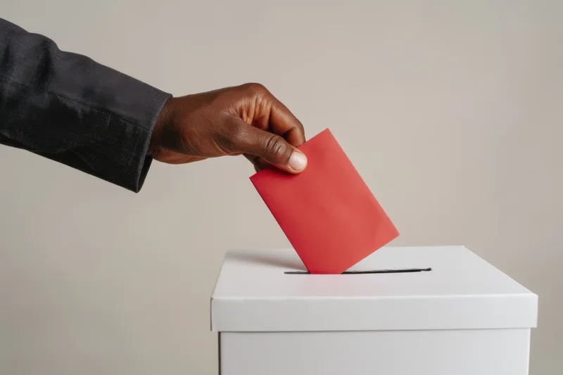 Hand in suit sleeve inserting red ballot into white ballot box
