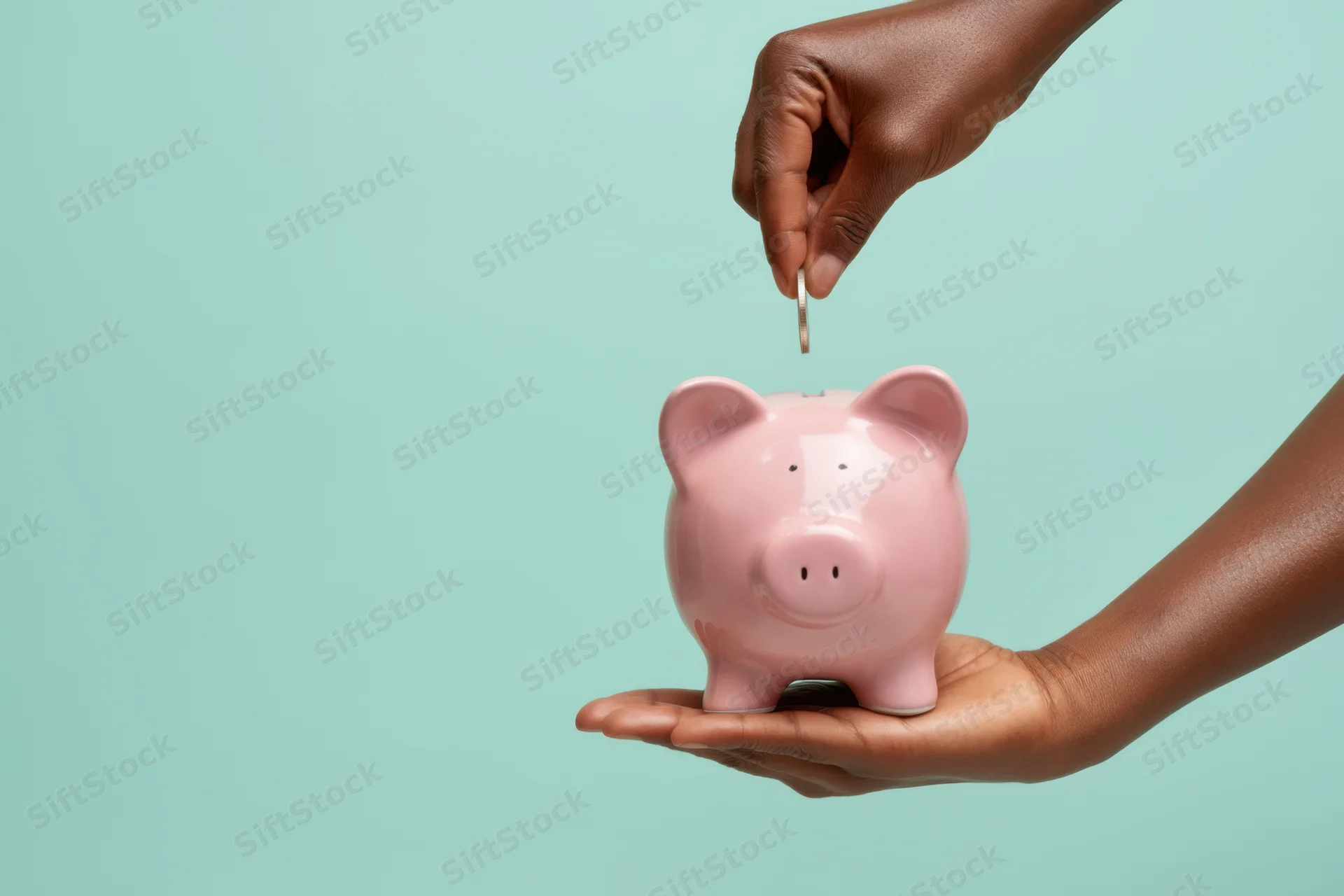 Black woman’s hand placing a coin into a pink piggy bank held in the other hand against a mint background