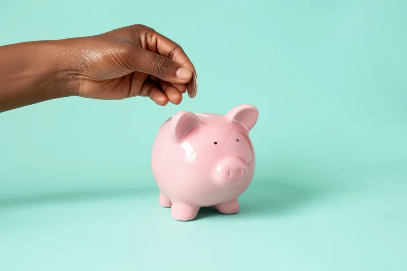Hand inserting a coin into a pink piggy bank on a turquoise background