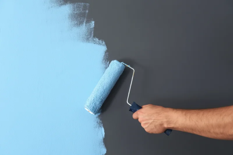 Hand holding paint roller applying light blue paint to gray wall during interior renovation