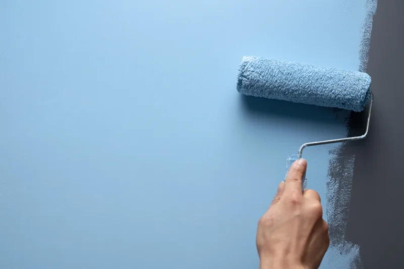 Hand holding paint roller applying light blue paint to wall