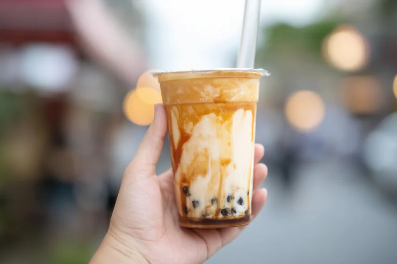 Hand holding a layered bubble tea drink with tapioca pearls and caramel sauce outdoors