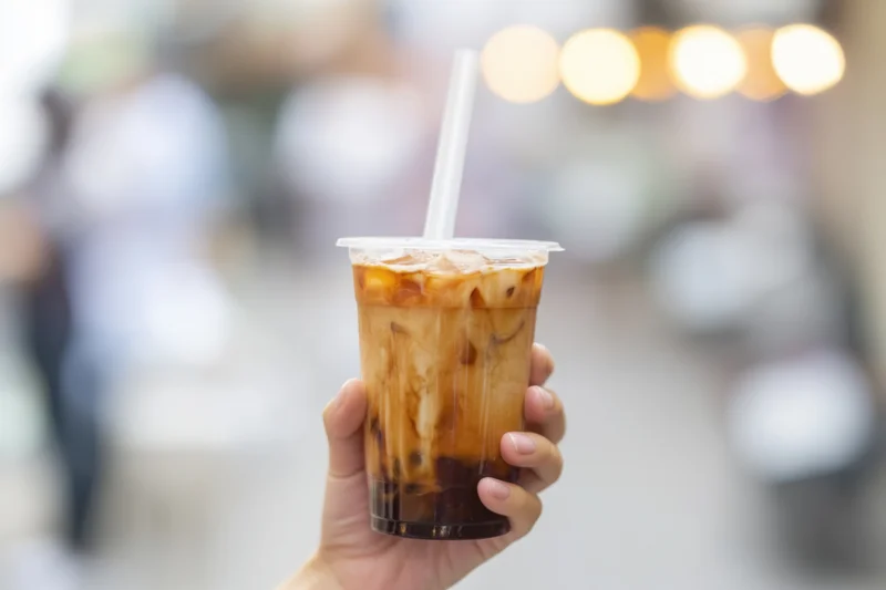 Hand holding a layered iced bubble tea with tapioca pearls in a plastic cup against a blurred urban street background