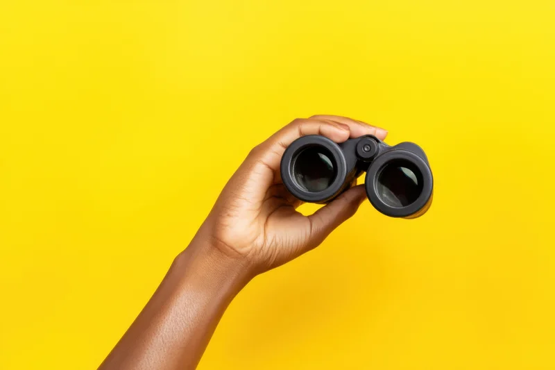 Hand holding binoculars against vibrant yellow background