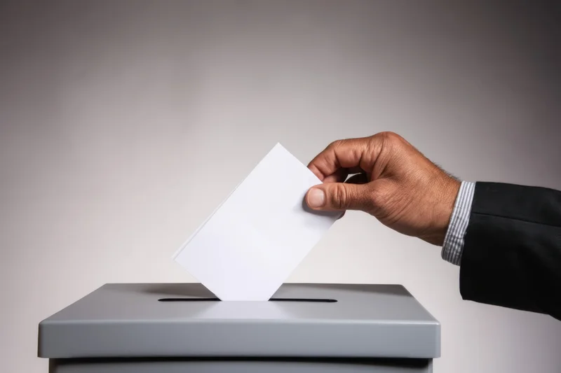 Hand in suit inserting blank white ballot into gray ballot box