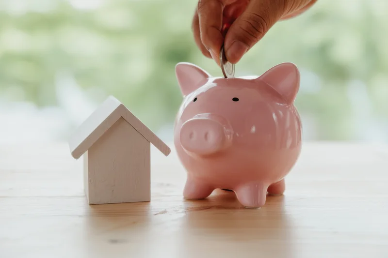 Hand inserting coin into pink piggy bank beside wooden house model on wooden table