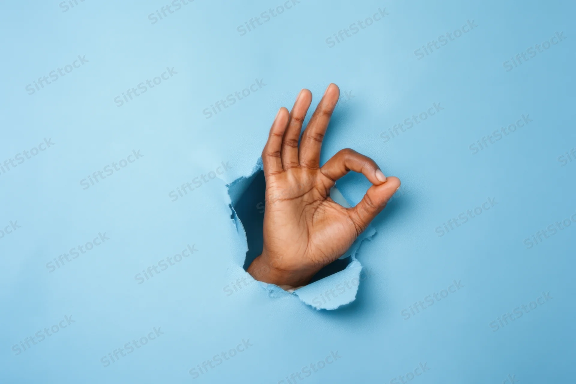 Hand making okay gesture emerging from torn blue paper