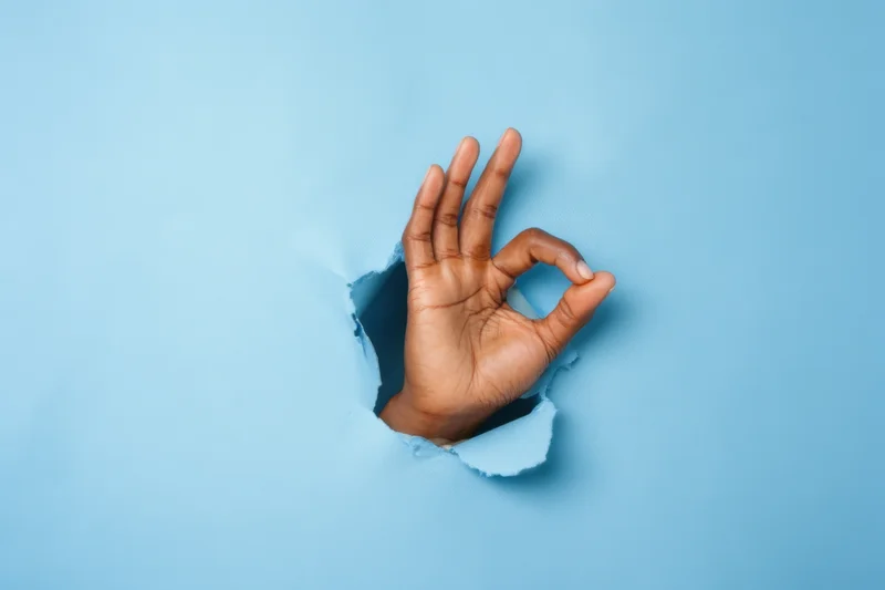 Hand making okay gesture emerging from torn blue paper
