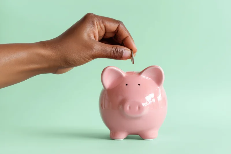 Hand placing coin into pink piggy bank against mint green background