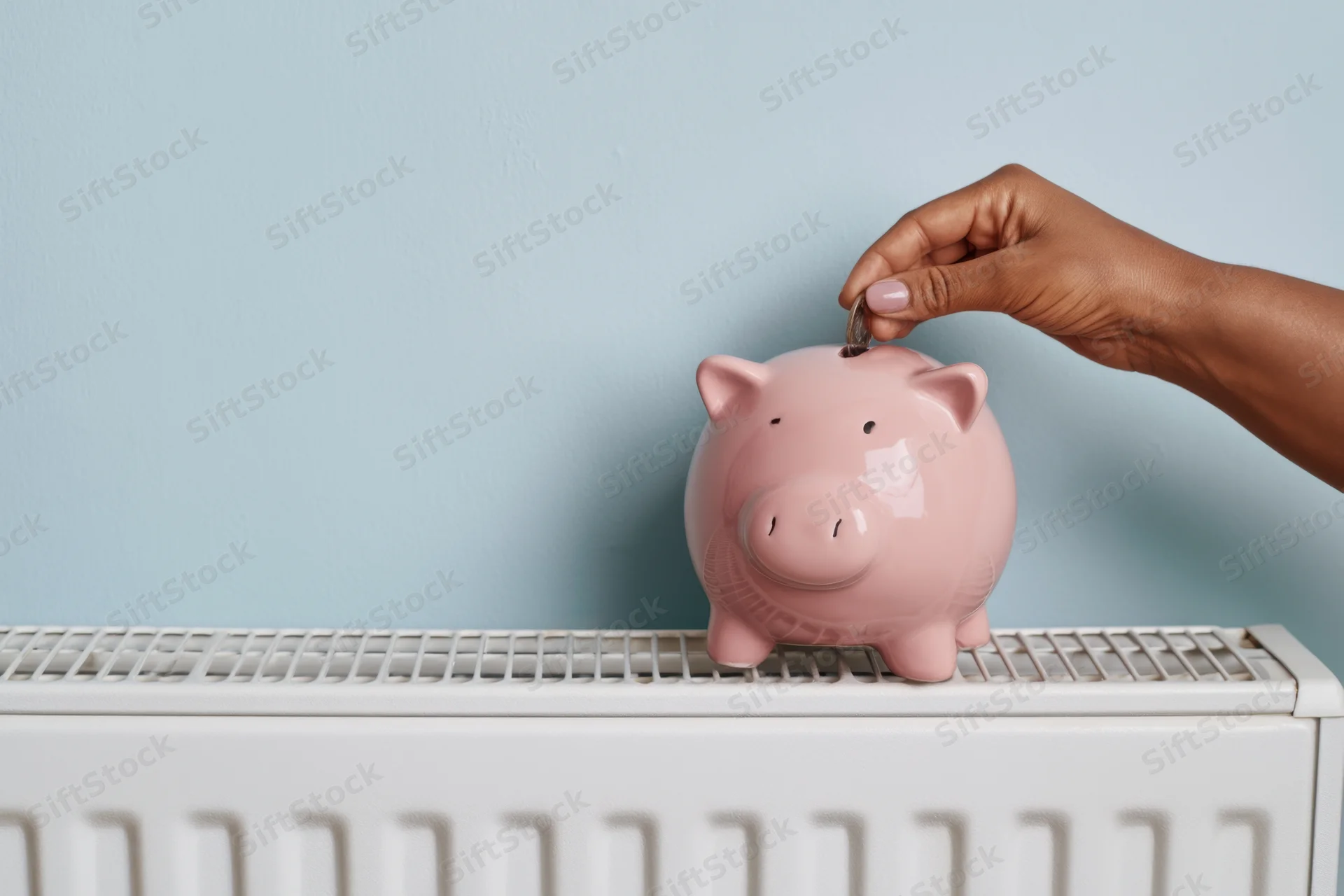 Hand placing coin into pink piggy bank on white radiator against blue wall