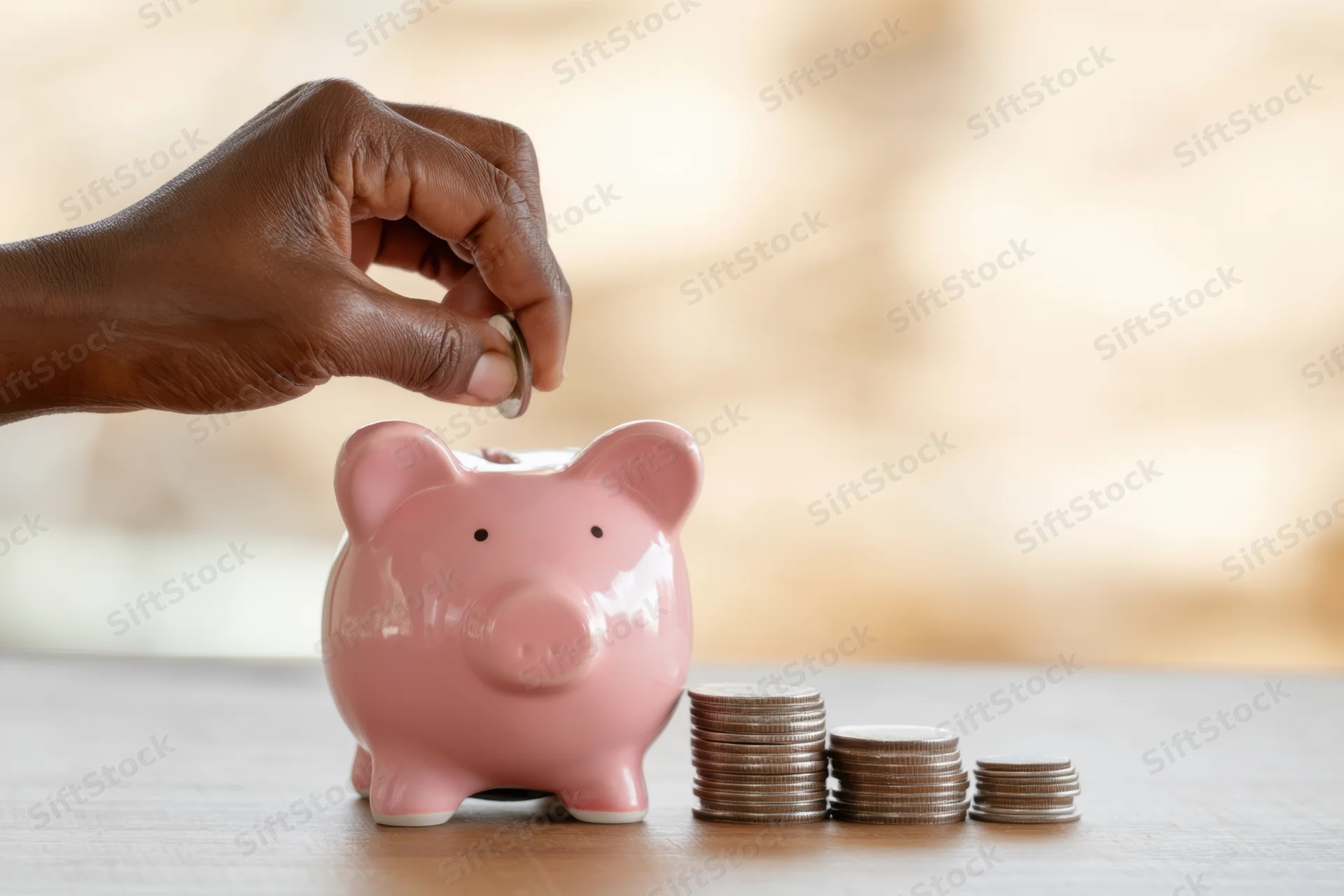 Hand placing a coin into a pink piggy bank on a wooden table beside stacks of coins arranged in ascending order