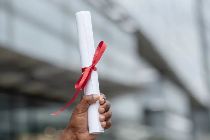 Hand holding rolled diploma tied with red ribbon against blurred background