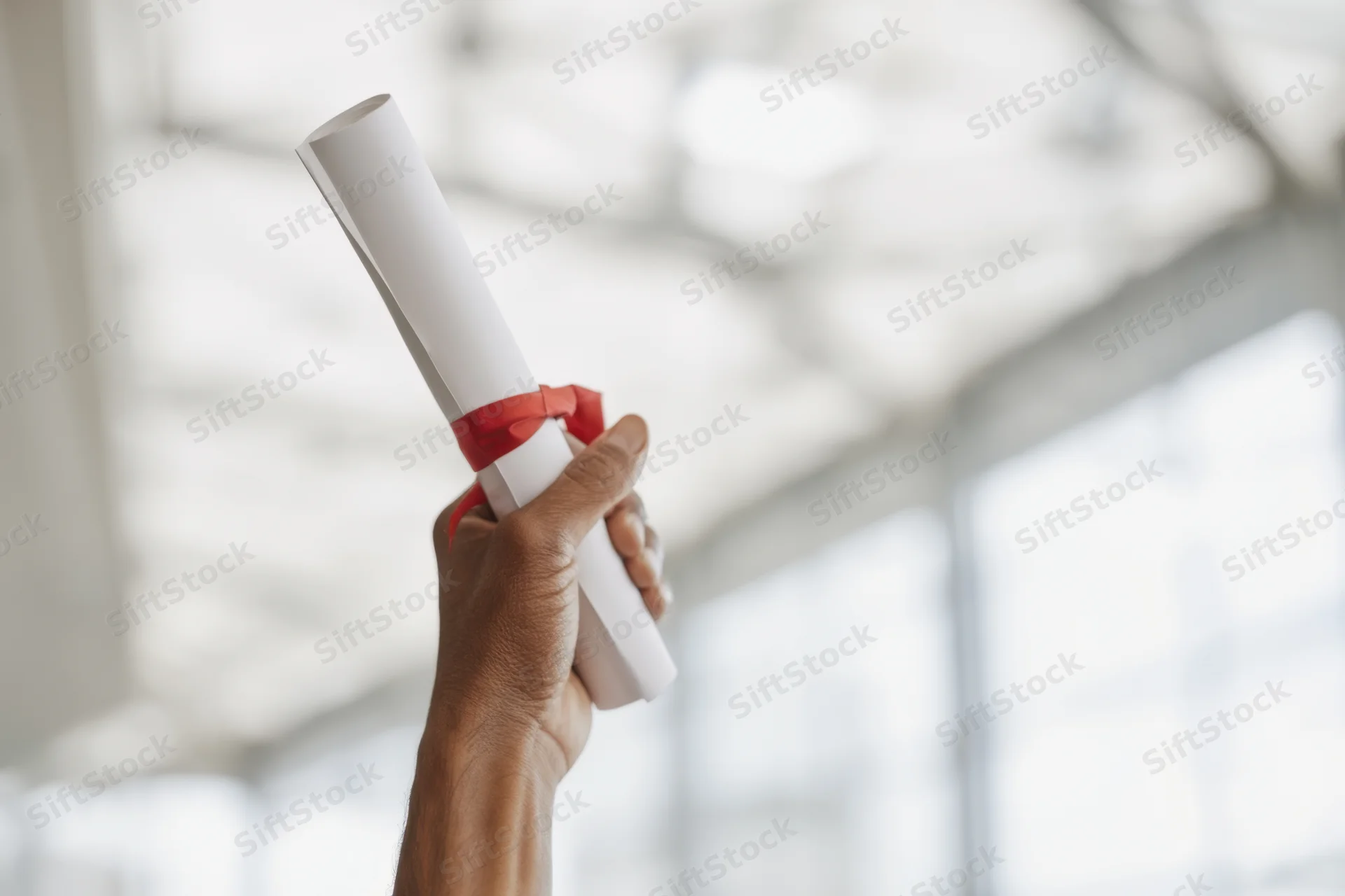 Hand holding up a rolled diploma tied with red ribbon in a bright modern interior