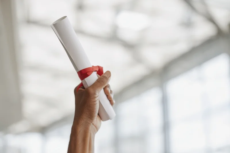 Hand holding up a rolled diploma tied with red ribbon in a bright modern interior