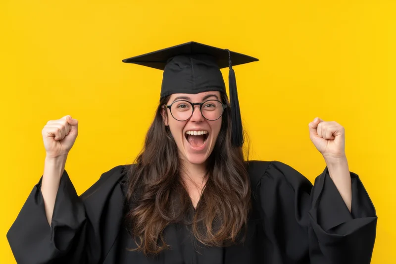 Joyful graduate in black cap and gown celebrating with fists raised against yellow background