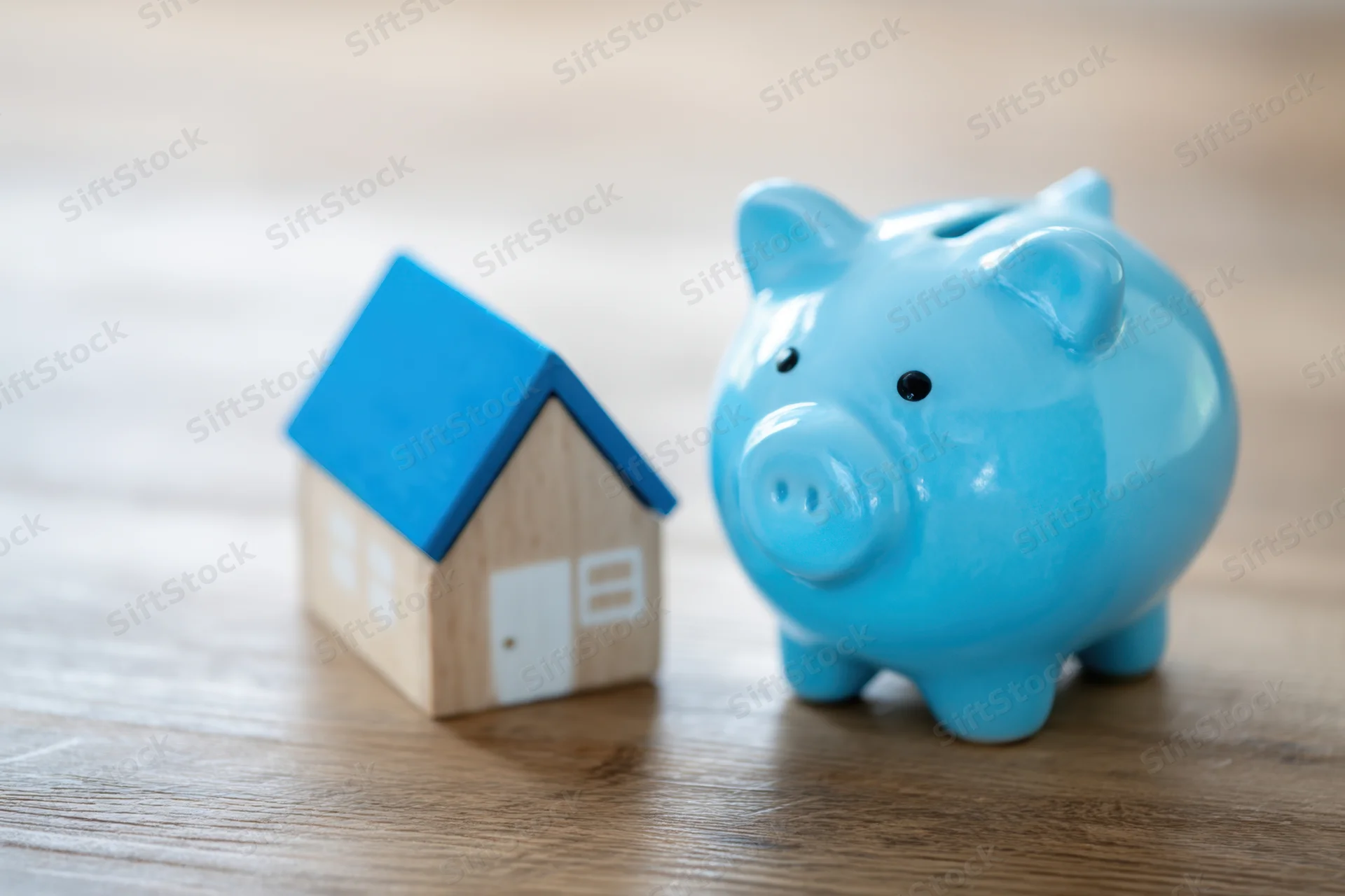 Light blue ceramic piggy bank next to a miniature wooden house on a wooden table