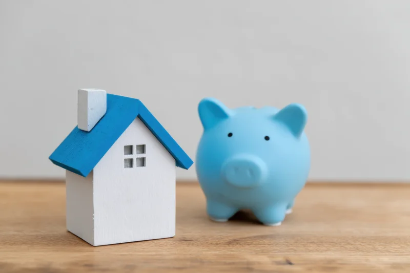 Light blue piggy bank and white wooden house model placed side by side on wooden surface