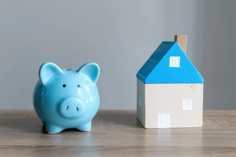 Light blue ceramic piggy bank beside a miniature wooden house on a wooden surface