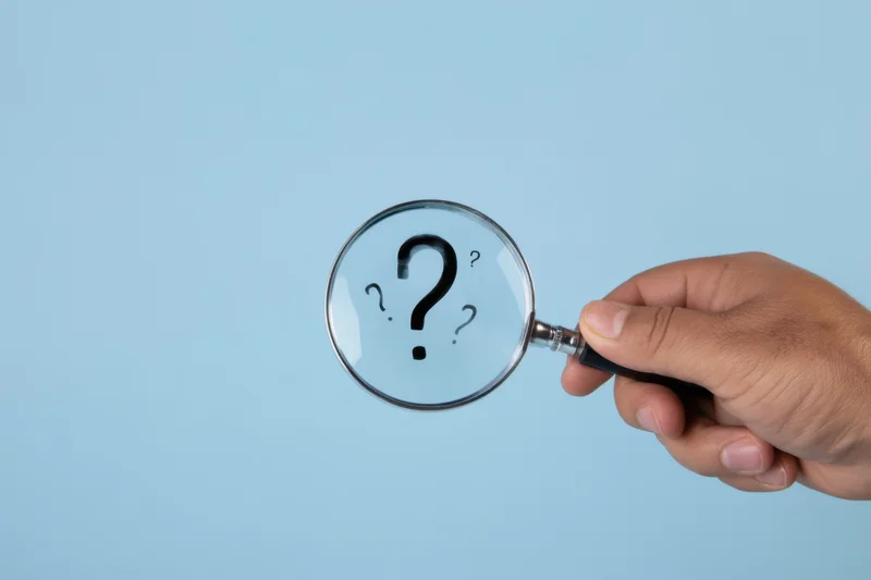 Hand holding magnifying glass focusing on large question mark against blue background