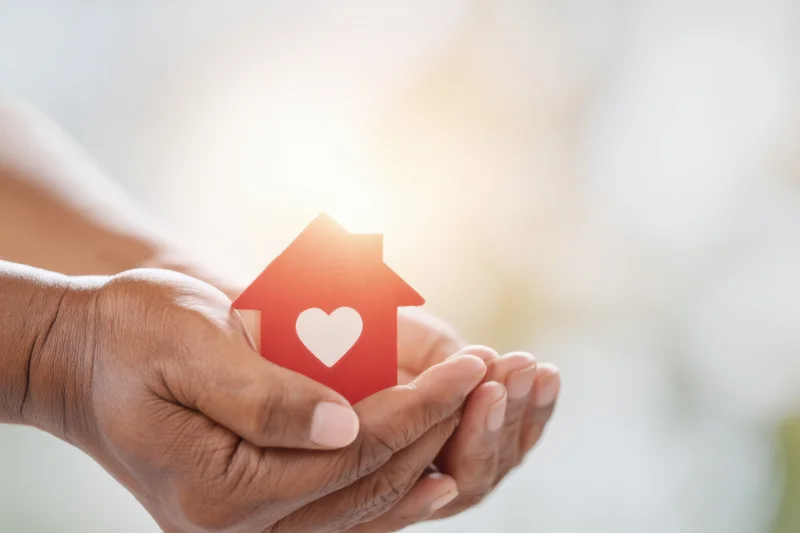 Hands cradling a small red paper house with a white heart cutout against a soft blurred background