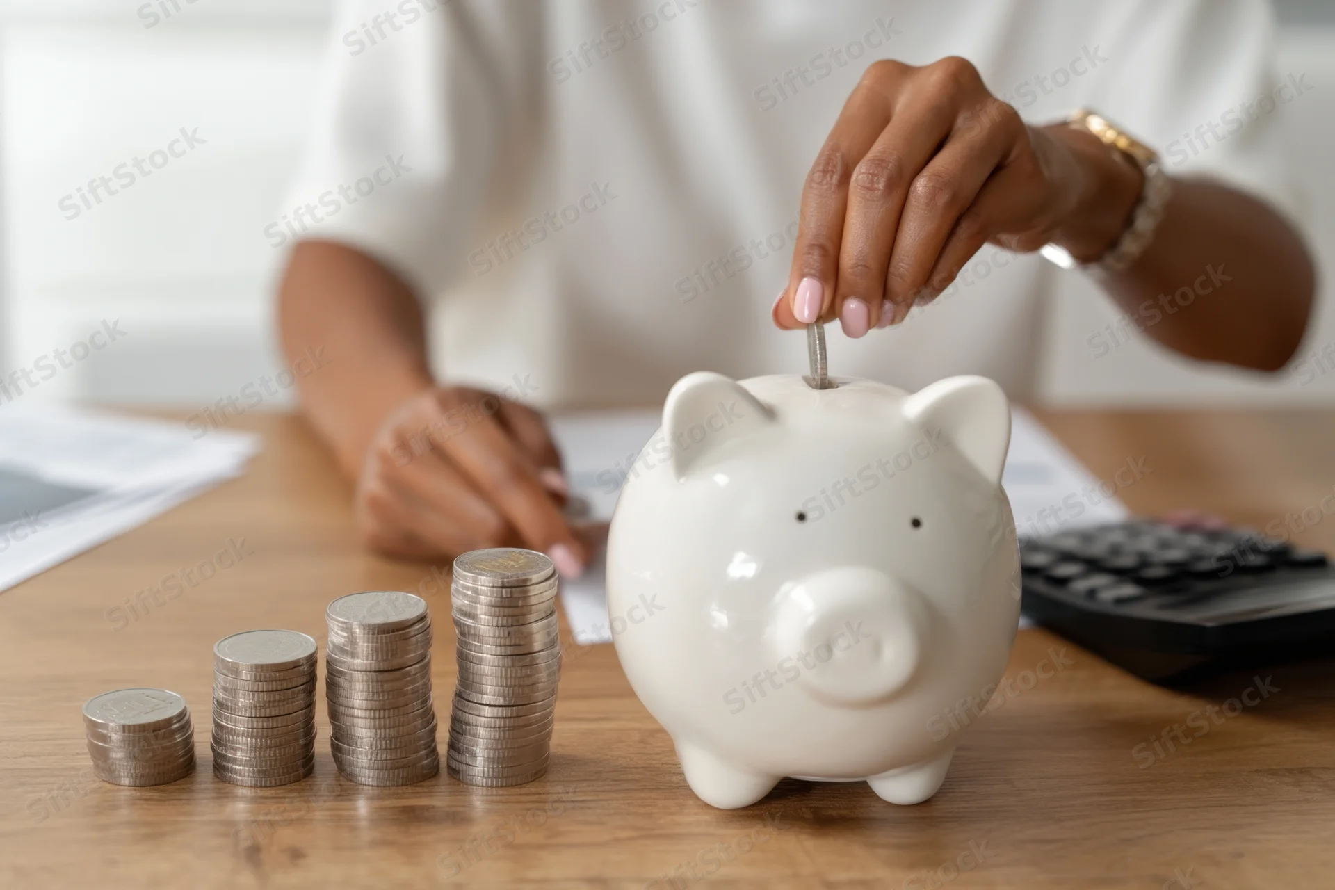 Woman placing coin into white piggy bank on wooden desk with stacks of coins and calculator nearby