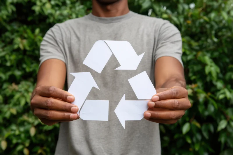 Man holding white paper cutout of recycling symbol in front of green foliage