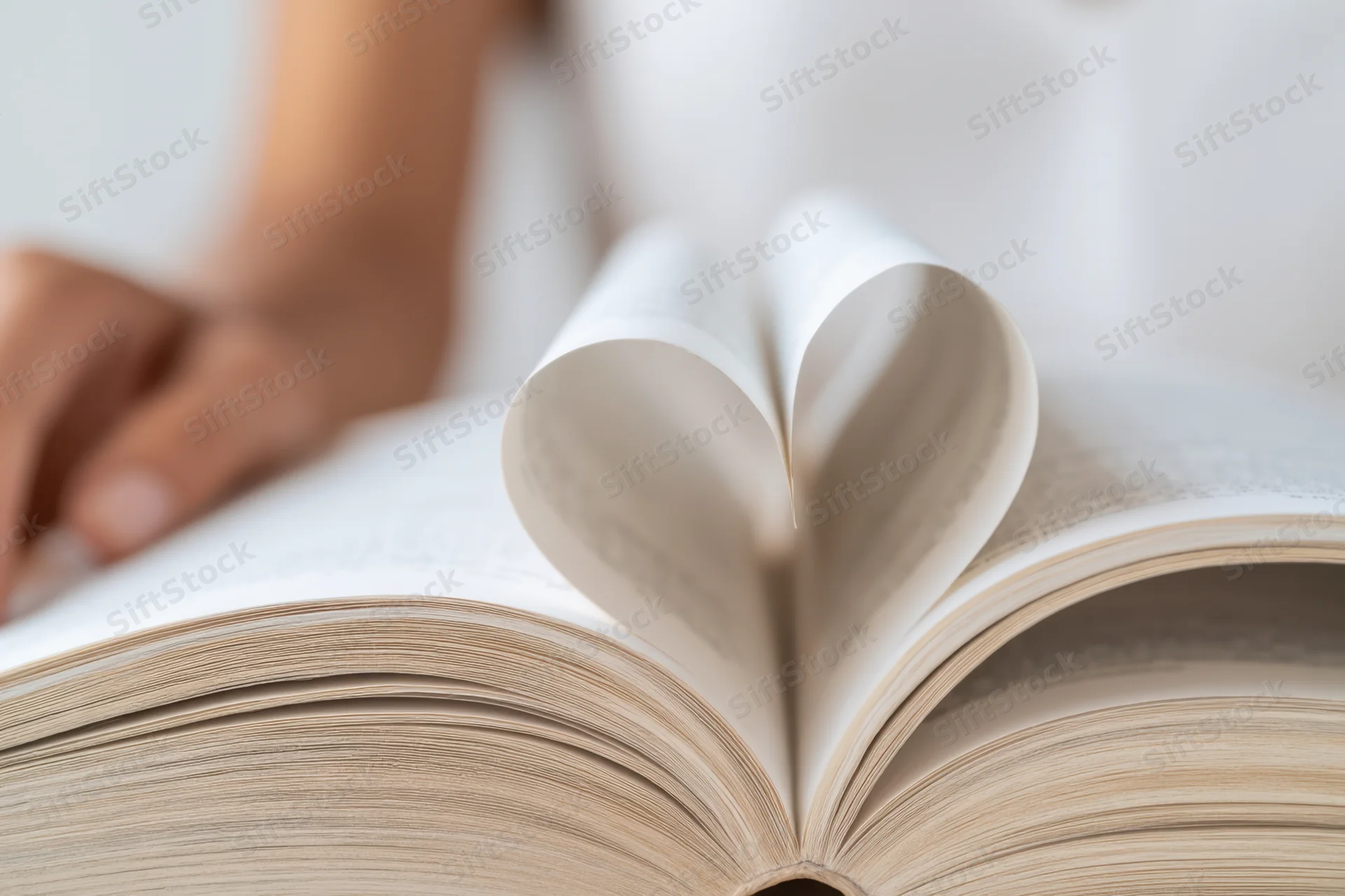 Open book with pages curled into heart shape, hand resting nearby on white surface