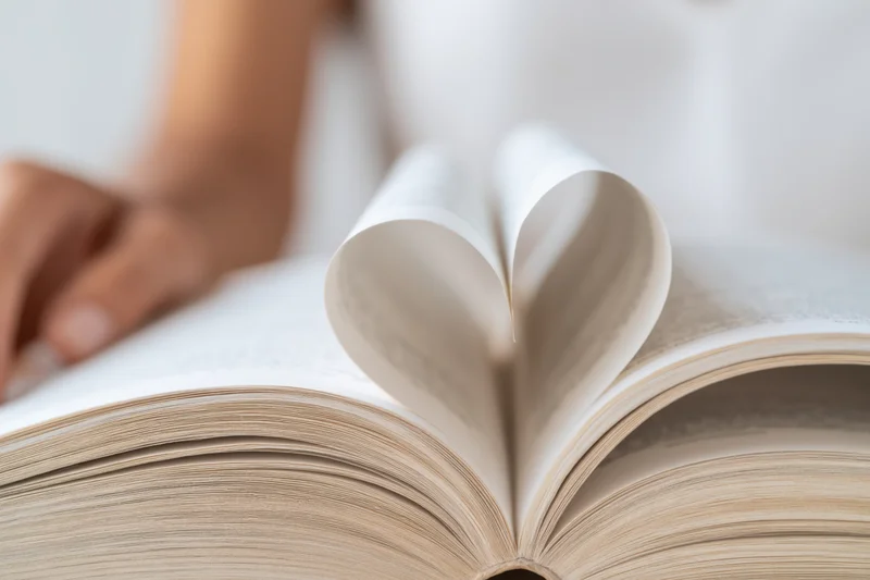 Open book with pages curled into heart shape, hand resting nearby on white surface