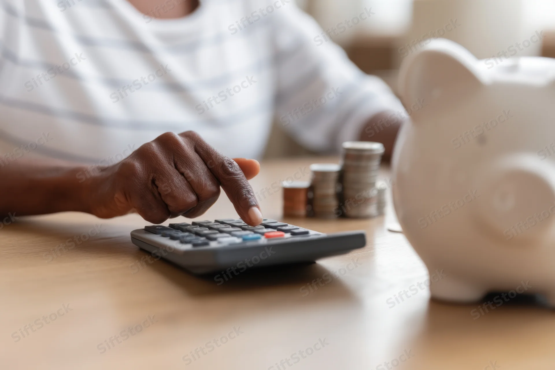 Person using calculator with coins stacked nearby and piggybank on wooden table