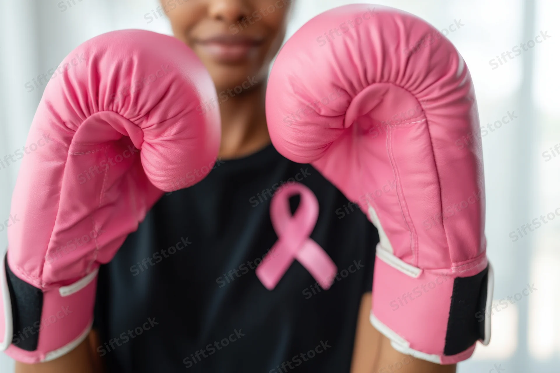 Woman wearing pink boxing gloves and black shirt with pink ribbon, smiling slightly with fists raised in front of her face