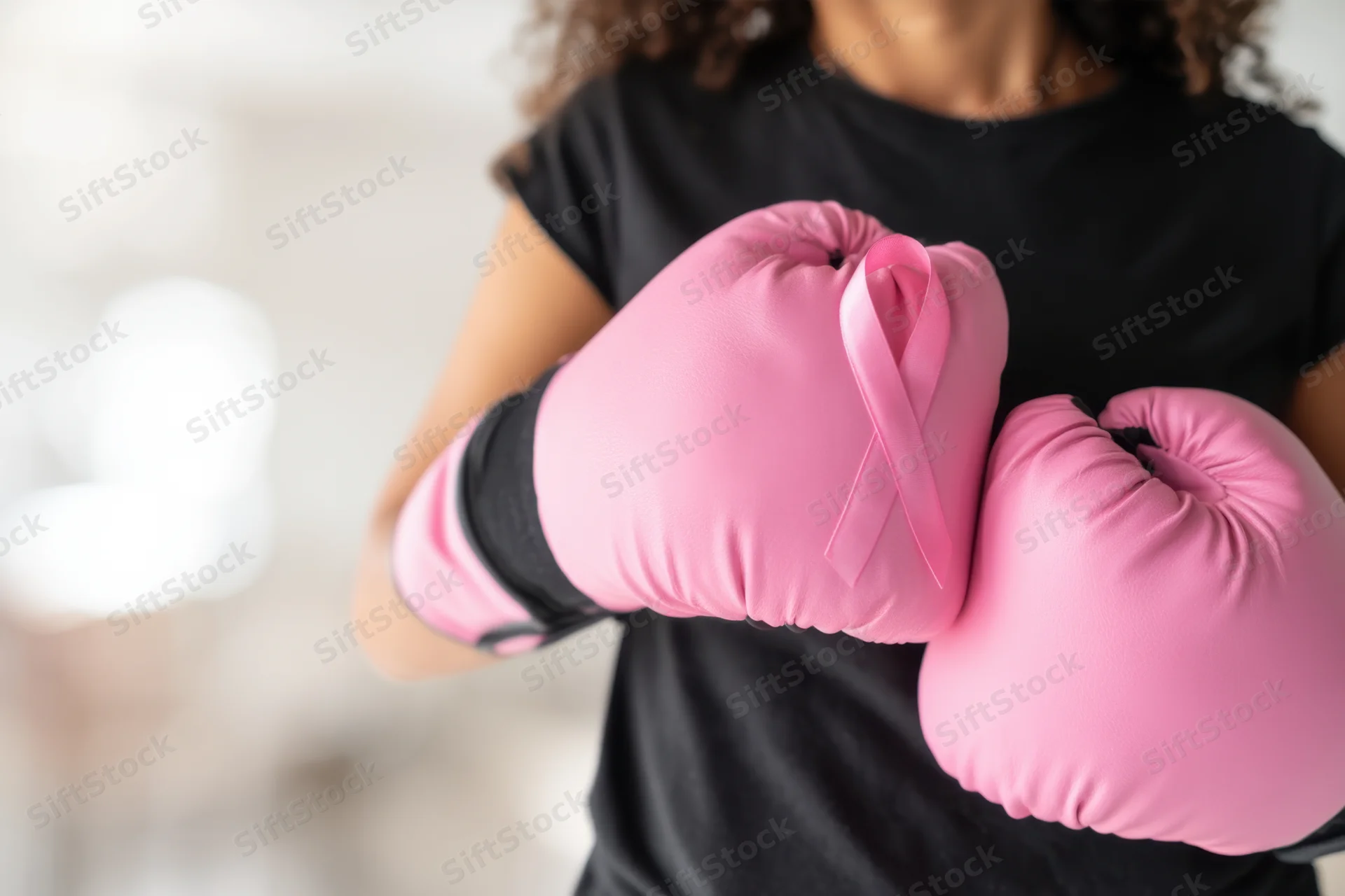 Woman wearing pink boxing gloves with breast cancer awareness ribbon, indoors in black shirt