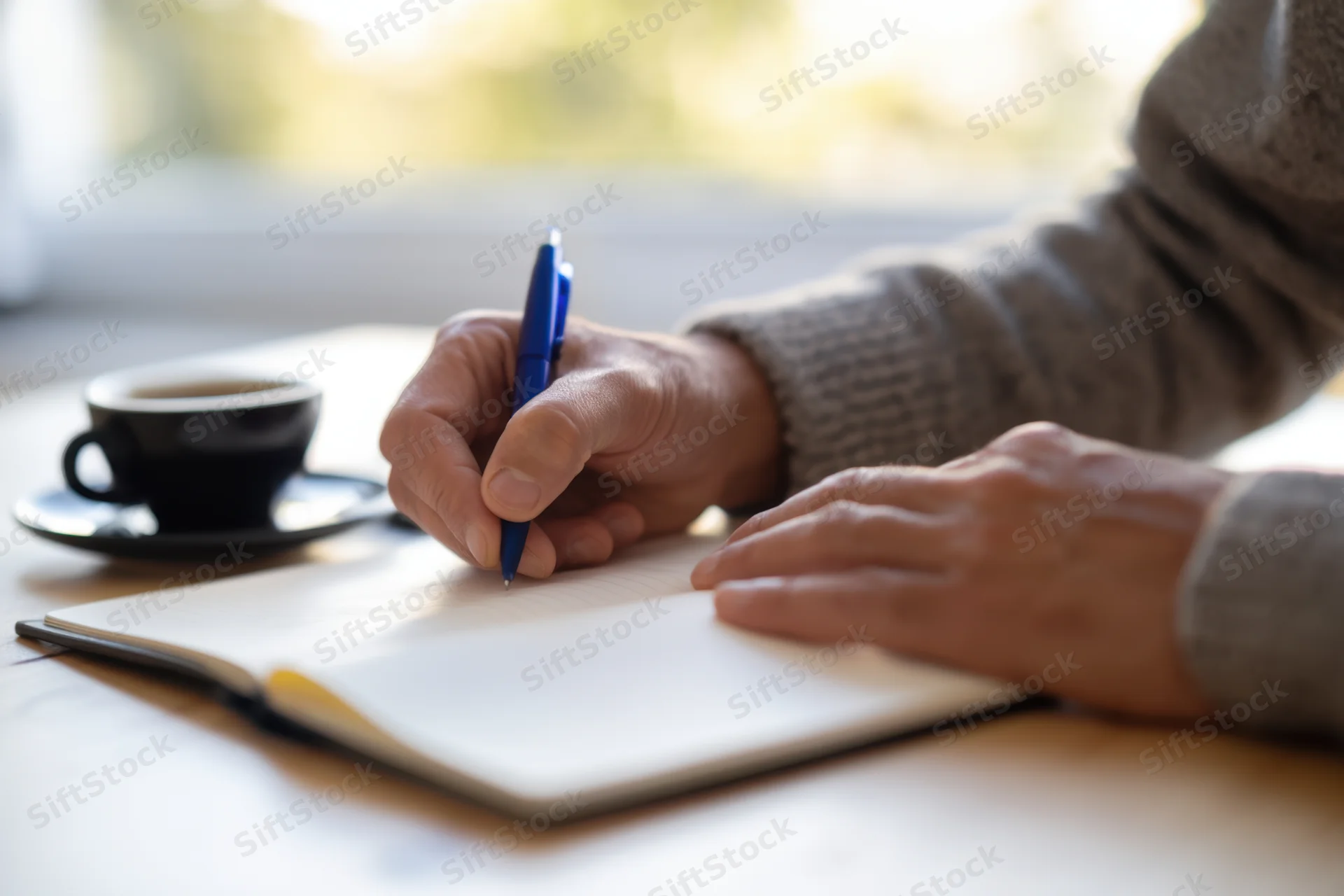 Person writing in notebook with blue pen beside coffee cup on wooden table