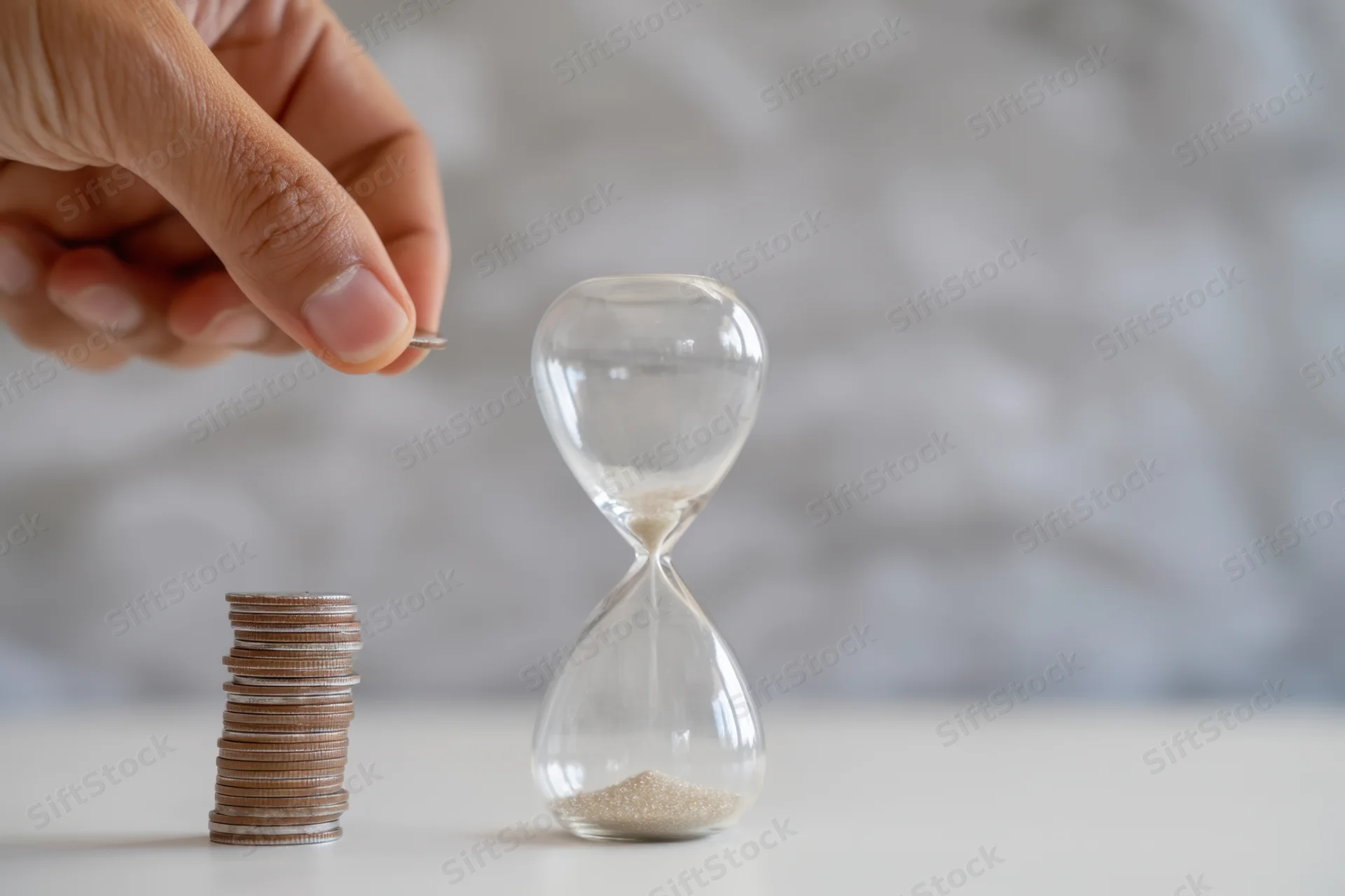 Hand placing coin on growing stack beside hourglass on white surface